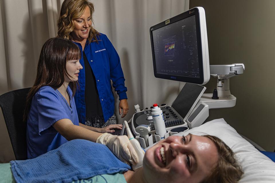 A student does an ultrasound on another student's neck while the professor stands by watching her work during scan lab.