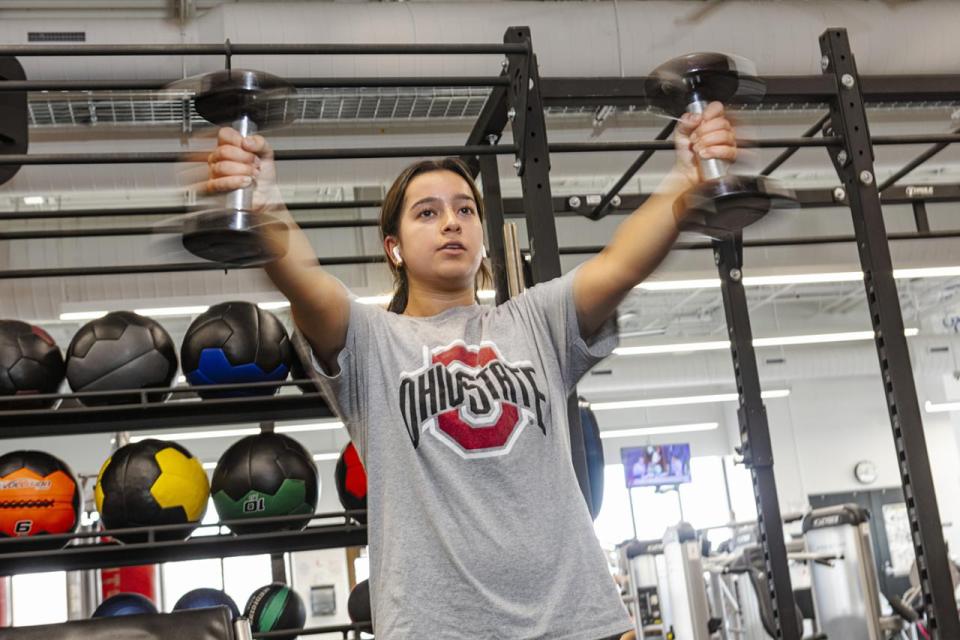 A student in a weight room holds dumbbells in her hands outstretched in front of her in motion.