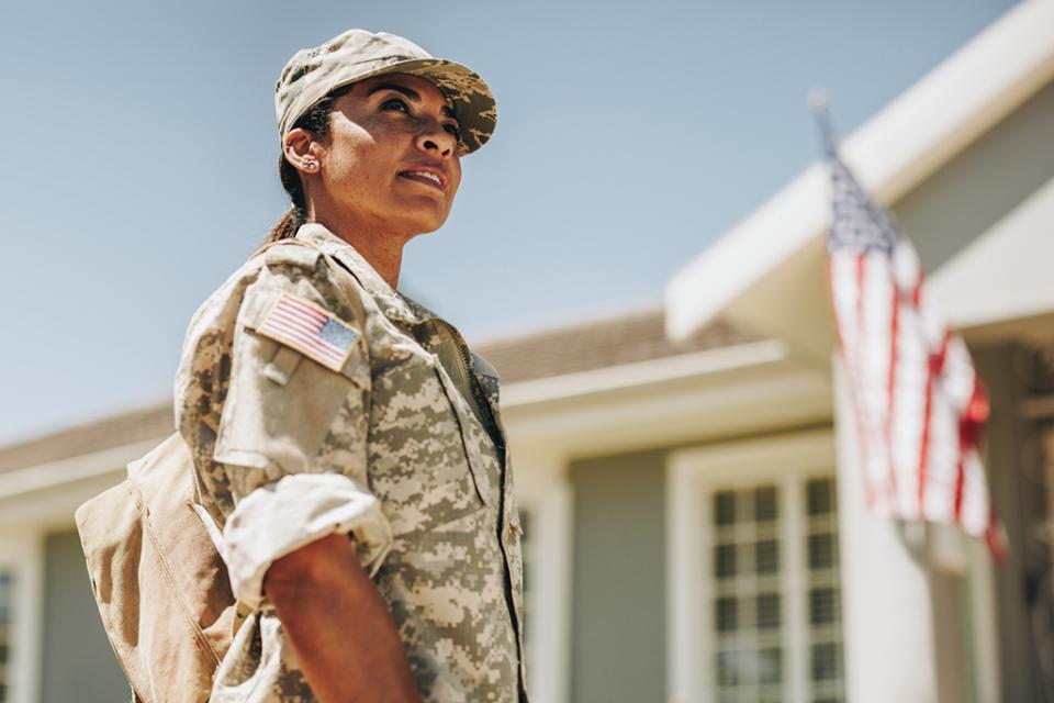 A person in camouflage military attire stands in front of a building with an American flag displayed.