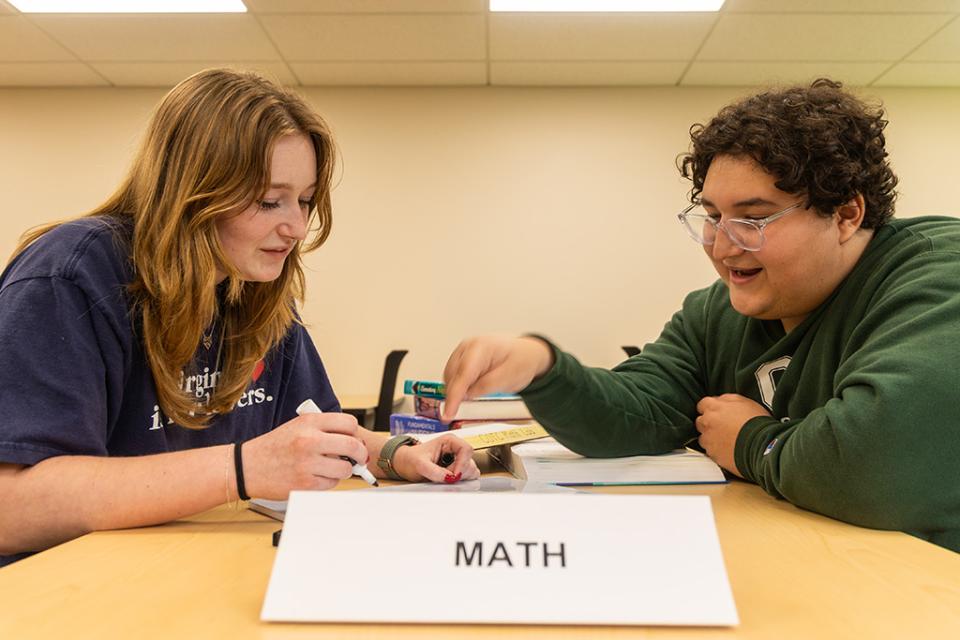 A student and a tutor sit a a table labeled "Math" working together on a dry erase board.
