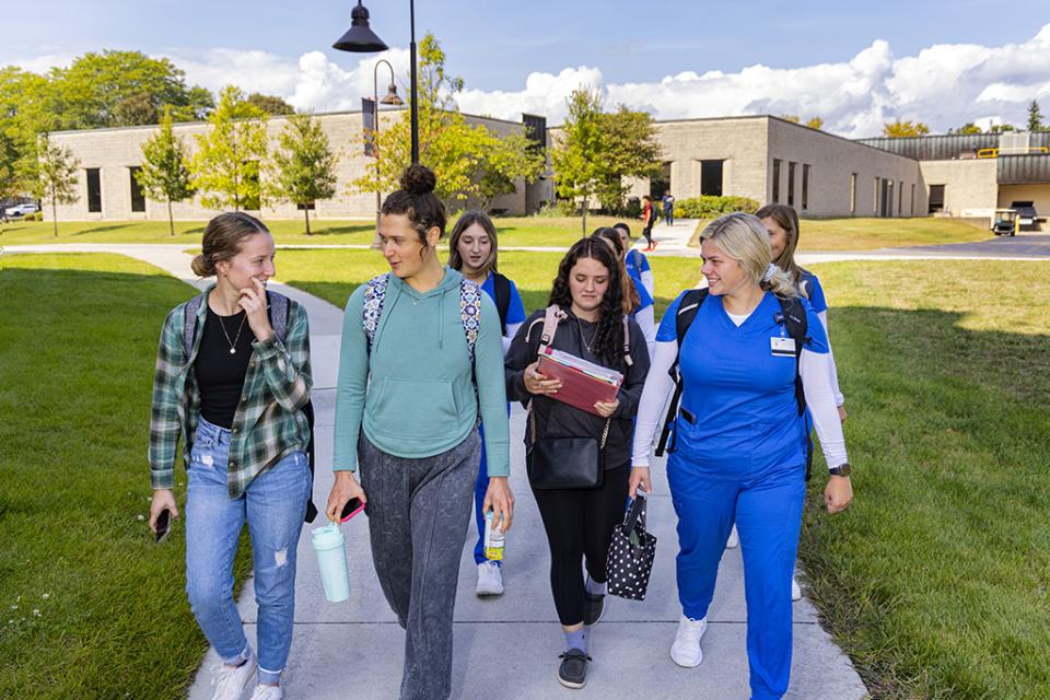 A group of students, some wearing nursing scrubs and some in plain clothes, walks on the sidewalk between buildings on campus.