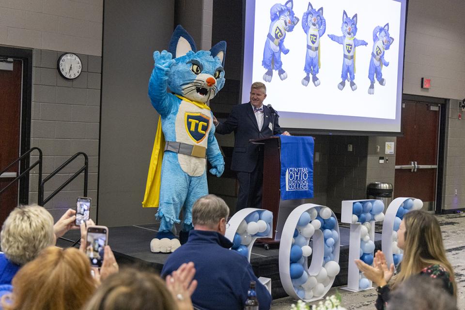 TC a blue cat with a yellow cape appears on stage next to COTC President John Berry in front of an audience.