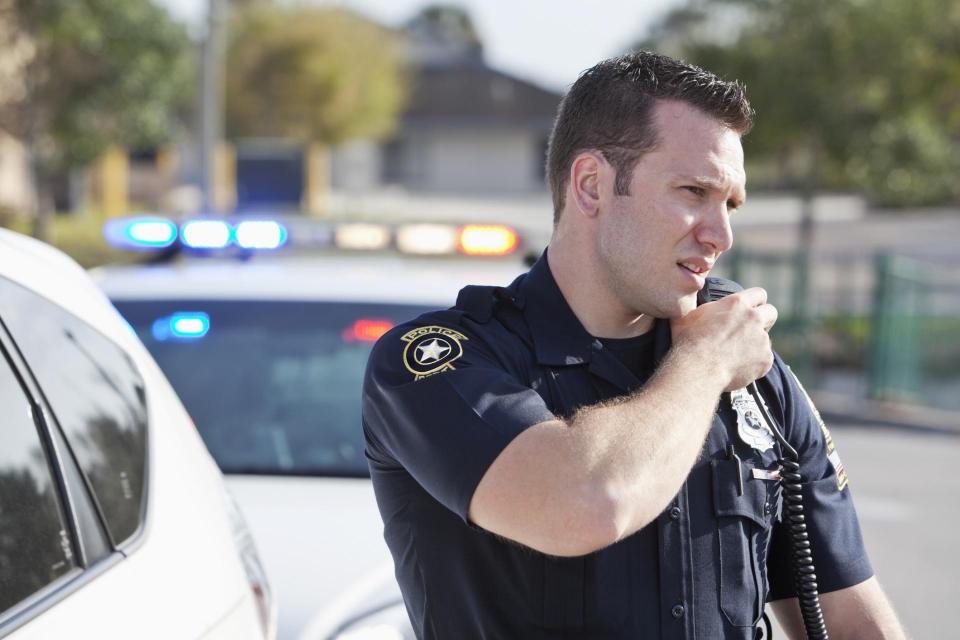 A uniformed police officer speaks into his walk-talkie while standing next to a police cruiser.