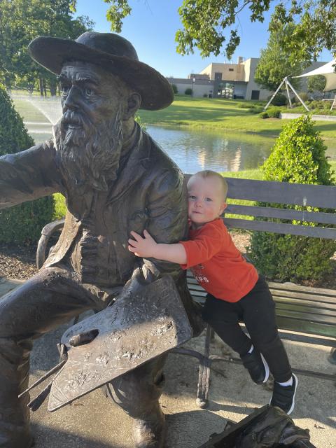 A toddler sits on a bronze bench statue holding the arm of a bronze Claude Monet statue.