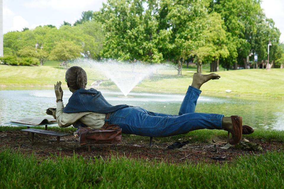 A statue of a girl lying on the grass gazing over the pond  with a book open in front of her.