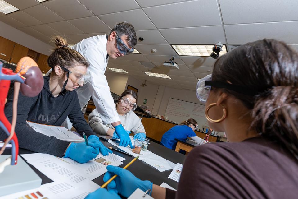 A professor wearing a lab coat and glasses stands between two students wearing protective equipment during an exercise in class.