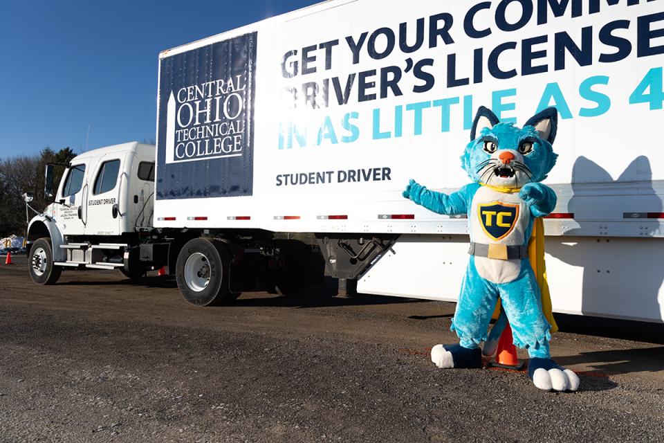 A blue cat mascot stands in front of a semi tractor trailer with the COTC logo on it.