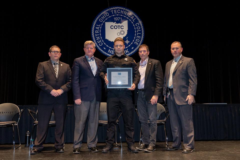 Srboljub Ruzic stands on stage holding his award certificate with Commander James Rease; COTC President John Berry, PhD; Rep. Troy Balderson; and Commander Adam Featherling.