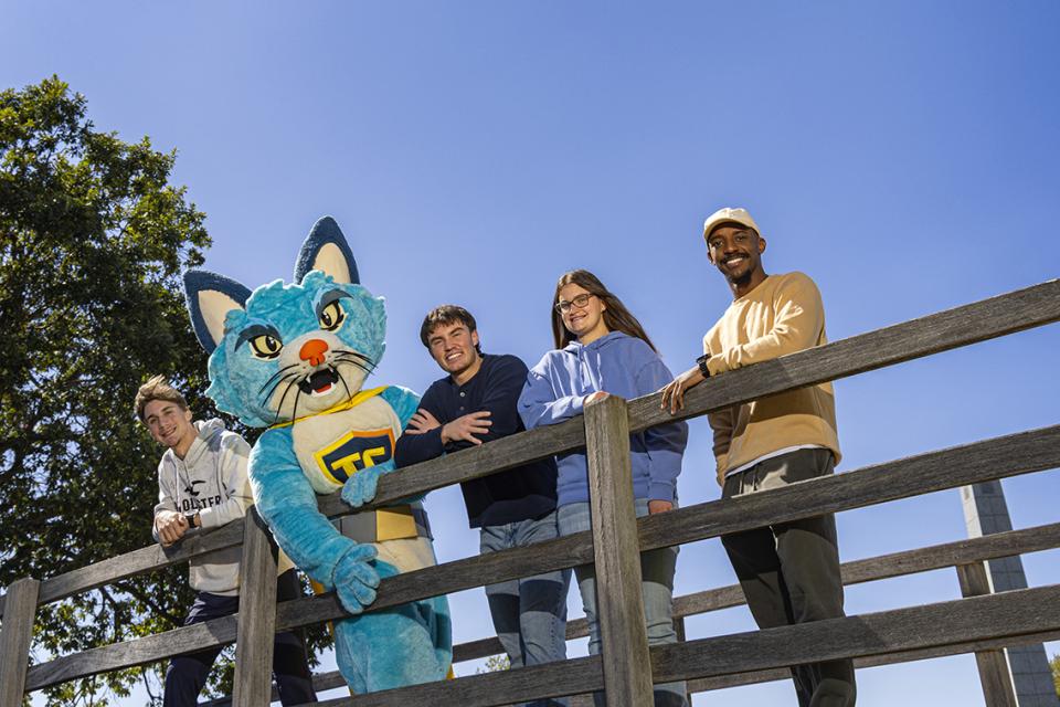 Four students gather around the COTC mascot, TC the cat, on a bridge at the Newark campus.