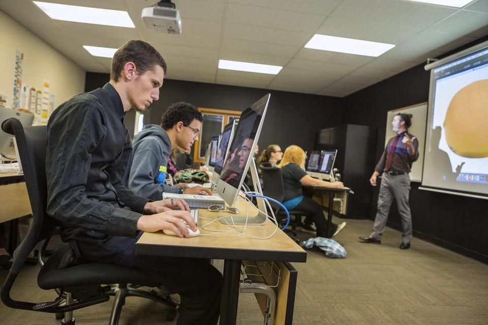 Students sit in front of a row of computers while a teacher stands at the front of the class. 