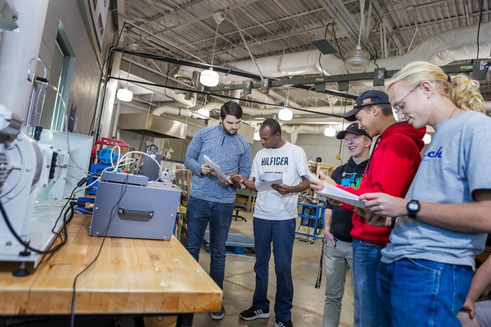 A group of students in the engineering lab  stands around equipment looking at a packet of paper in their hands.