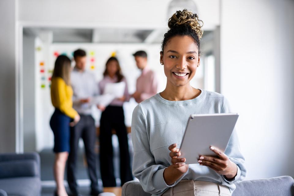 Portrait of a young woman holding a tablet in an office with a group of people behind her. 