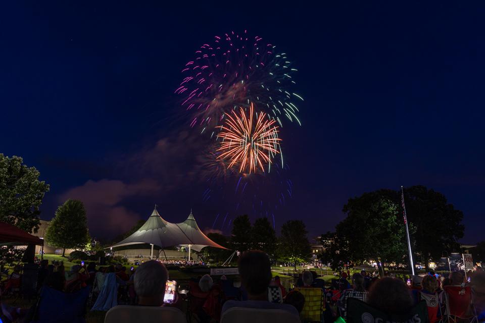 Fireworks explode over the campus while onlookers watch from the campus lawn.