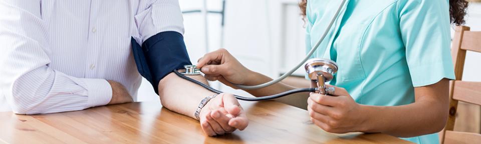 Female nurse taking blood pressure of elderly male, both sitting at table