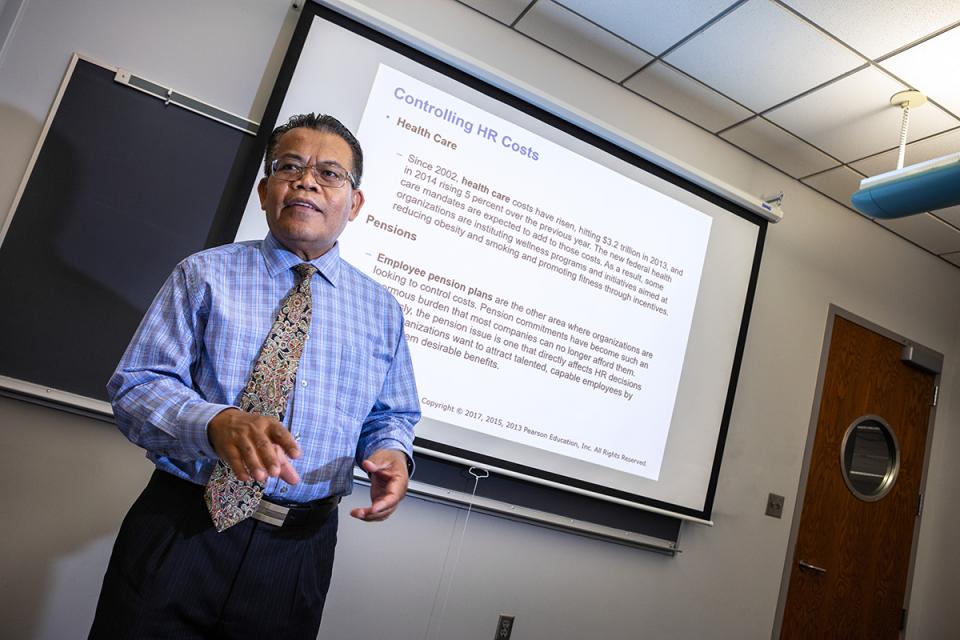 Professor stands at the front of a classroom in lecture looking toward students. 