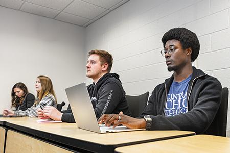 A student on a laptop sits at the end of a row of students seated at a desk.