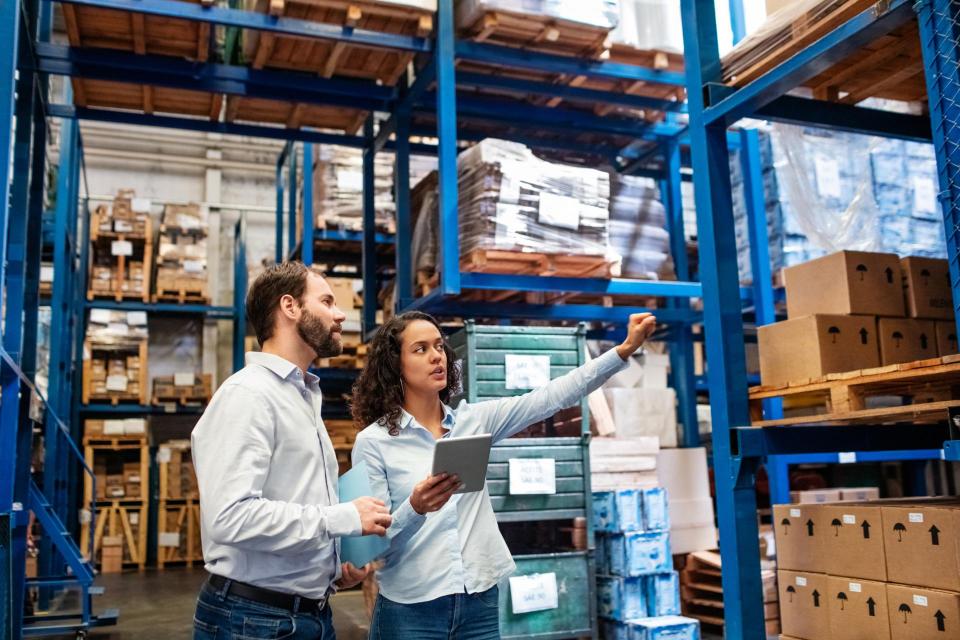 A man and woman look in the direction of her outward stretched hand in a warehouse that has stacks of boxes and product.